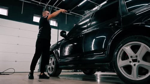 A male car wash employee applies car wash detergent to a black luxury car using spray gun