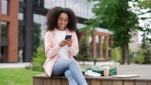 Smiling african american female student using mobile phone sitting on a bench in campus space near