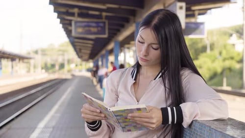 A Young Beautiful Caucasian Woman Reads a Book at a Train Station