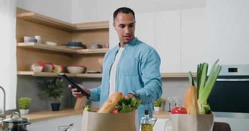 Man stand by kitchen table using smartphone search healthy recipes for balanced diet, ordering produ