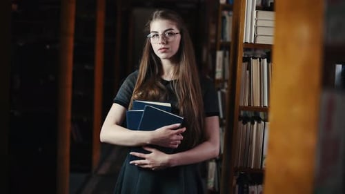 Brunette in eyewear stands and smiling in the library with books in hands