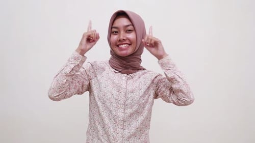 Happy young asian muslim girl pointing presenting above and showing thumbs up to the camera. Isolate