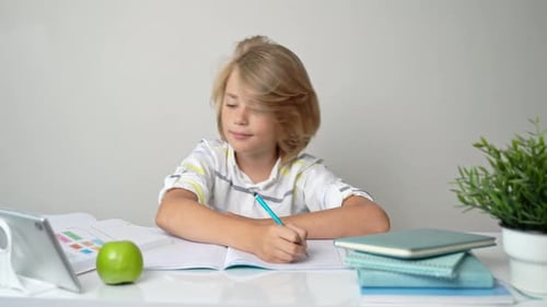 Middle School Smiling Student Boy Sitting at Desk Studying Writing Book Homework and Tablet at Class