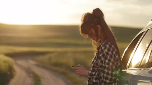 Young Woman Using Phone by Car at Sunset
