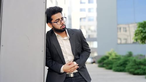 Young bearded businessman in a formal suit feels stomach pain standing on the street near an office