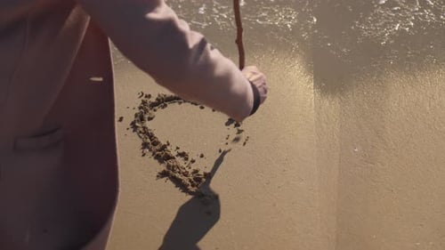 Junge Frau zeichnet am Strand ein Herz in Sand, bevor die Meereswelle es wegspült