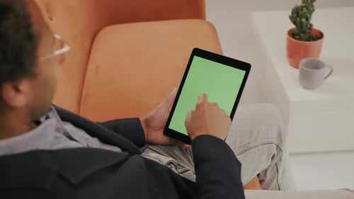 Man on sofa with tablet, zooming and tapping the green screen, watching and nodding.
