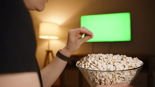 A woman eats popcorn in front of the television. Green screen. Dim and warm light from the lamp.
