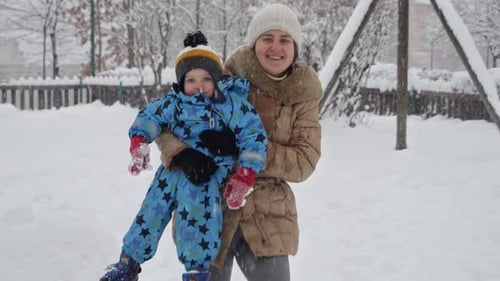 Portrait of happy smiling mother holding her toddler son and walking over snowdrifts on playground