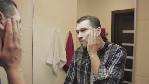 Junger Mann, der in den Spiegel schaut, berührt sein Gesicht, bevor er sich im Badezimmer rasiert, Zeitlupe