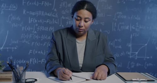 Portrait of African American Woman Scientist Writing in Notebook Sitting in Room with Chalkboard