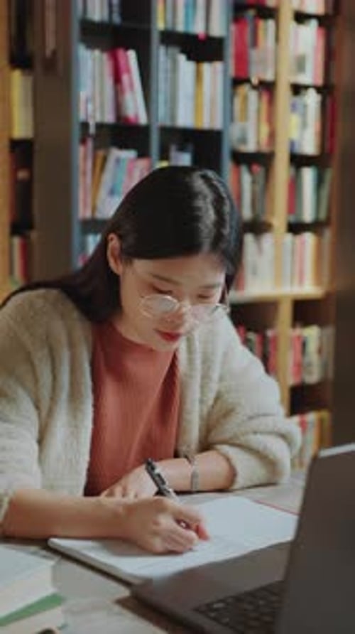 Asian Girl Reading from Laptop and Writing Notes at Desk in Library