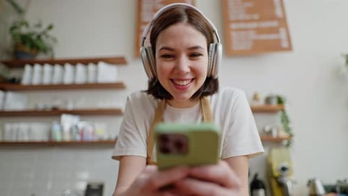 Happy Brunette Girl in White Wireless Headphones Tshirt and Apron Leans on the Counter and Listens