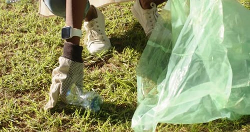 Mujer afroamericana que ve una botella en la hierba en cuclillas y coloca la botella en una bolsa verde para limpiarla
