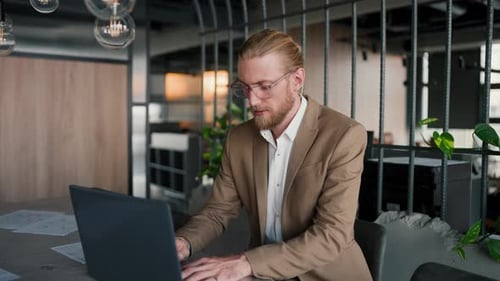 A Blond Guy in Glasses with a Beard in a Light Brown Suit Works at a Laptop and Then Looks at the