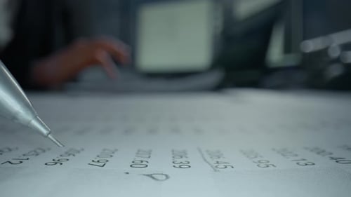 POV of Office Desk Closeup with Pencil Marking Numbers on Paper While Another Person Uses Calculator