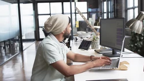 Young Asian man works on laptop in a modern business office setting, wearing headphones and a beanie