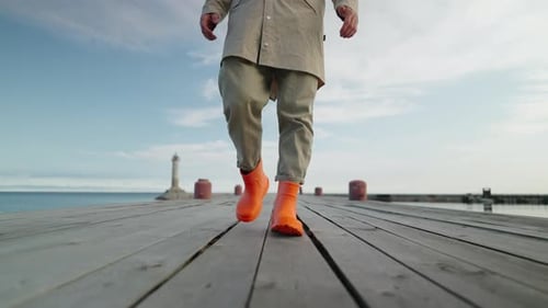 Fisherman In Bright Orange Rubber Boots Walking On Old Wooden Pier In Morning View On Male Legs