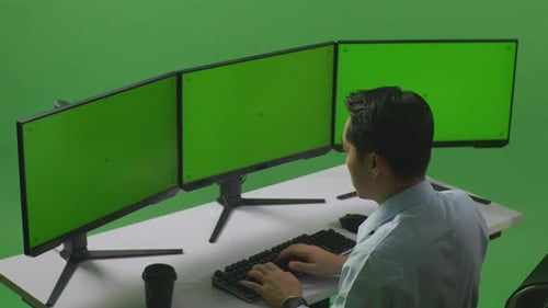 Side View Of Asian Man Working With Mock Up Multiple Computer Monitor In Green Screen Studio