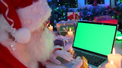 Santa Claus having video chat at laptop with green chromakey screen, bearded grandfather with gift