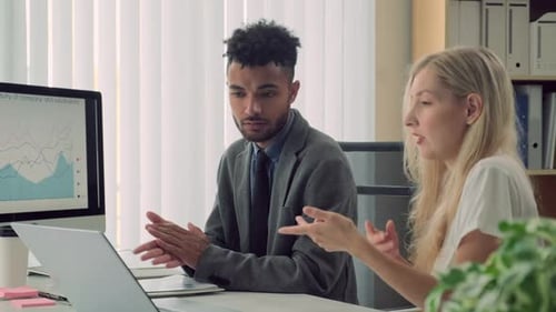 Two Office Workers Discussing Details of Project Sitting at Table with Laptop