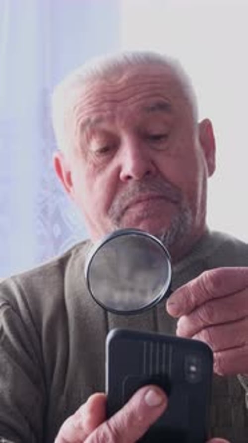 Senior Man Examines Smartphone with Magnifying Glass Indoors