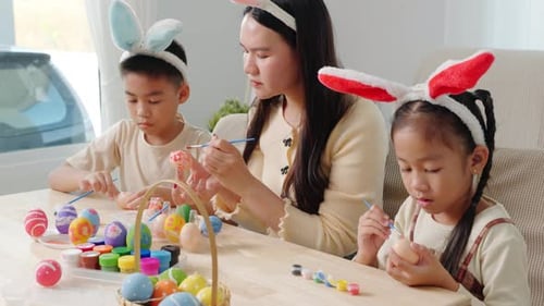 A Mother and Kids with Bunny Ears Happily Paint Easter Eggs at a Table Near a Festive Basket
