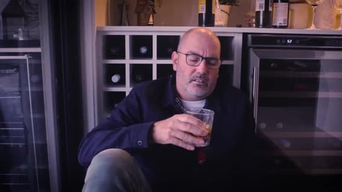 A disappointed man sits on the floor, banging his head against a wall - wine rack while drinking spi