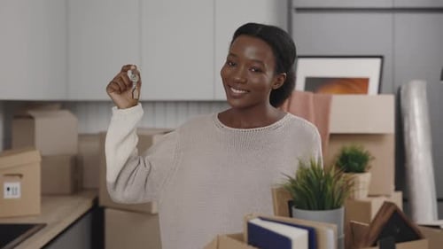 Closeup of African American Female Holding House Key Looking at Camera and Smiling Indoors in Light