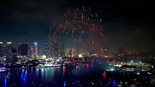 Elevated drone shot of fireworks bursting in brilliant patterns, mirrored in the bay with city