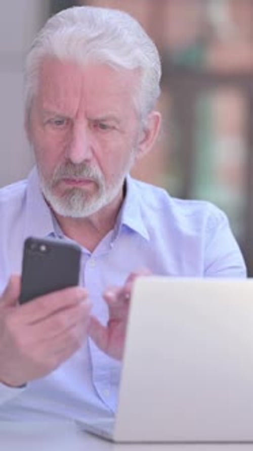 Focused Senior Man Using Phone and Laptop Indoors