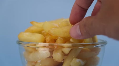 Shot of hand picking up fresh french fries in glass bowl on blue background.