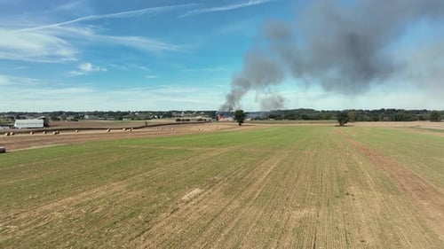 Drone flight over farmland field and burning American farmstead in background. Rising dark smoke