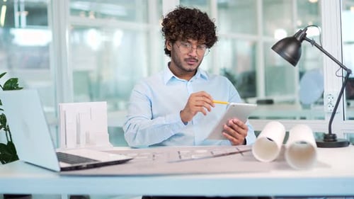 Young handsome architect draws a project on paper using a tablet while sitting at a workplace at a d