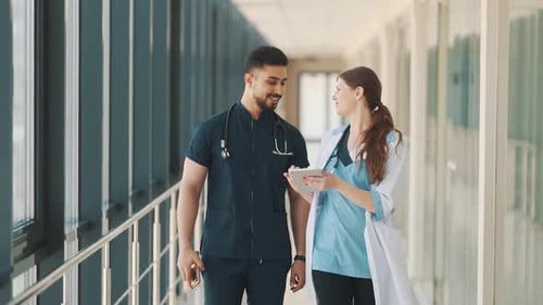 A doctor and a nurse are talking while walking along a hospital corridor with a tablet