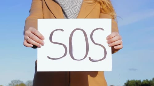 Woman hands showing SOS poster sign towards camera in front of clear blue sky, wearing yellow winter