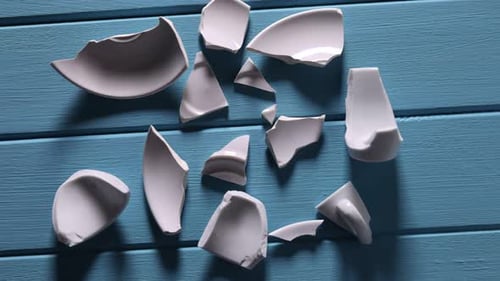 Pieces of broken saucer and cup on light blue wooden table, flat lay. Camera moving forward