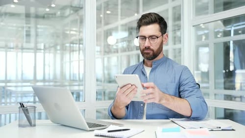 Confident businessman is using digital tablet sitting at workplace in business office. Handsome