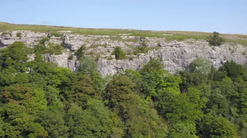 Drone footage flying sideways and ascending up and over a steep, tree lined limestone crag revealing