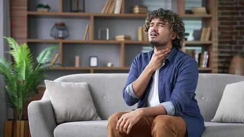 Man Sitting on Couch Holding Sore Throat in Pain