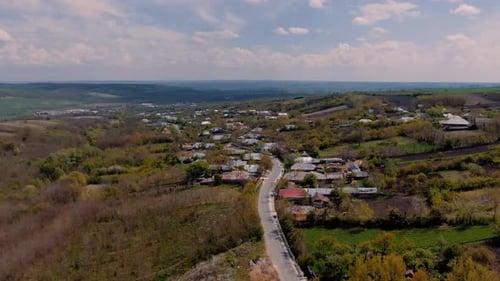 Aerial view over an old village road and houses, agricultural fields, surrounding hills, partly clou