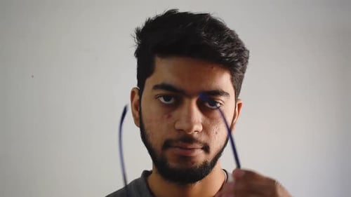 junger erwachsener Mann, der die Brille abnimmt, zwinkert und lächelt allmählich. Nahaufnahme des jungen Ha