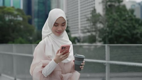 Attraktive junge muslimische Frau, die in der Kaffeepause draußen mit Hijab läuft und ihr Handy benutzt