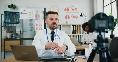Portrait of man doctor recording video on camera for internet patients sitting at his workplace