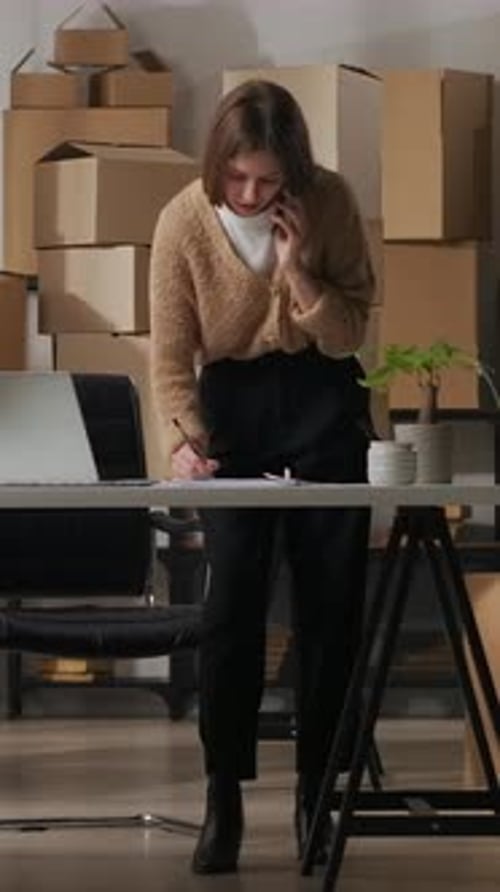 Focused Small Business Owner Managing Orders on Phone and Laptop in Home Office Workspace Surrounded