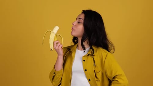 Smiling Beautiful Woman Enjoying While Eating Banana Posing in Studio Isolated on Yellow Background