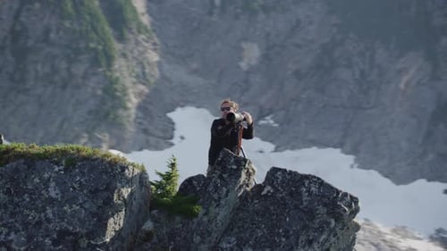 Photographer double checks camera view on mountain ridge, medium tracking helicopter counterclockwis