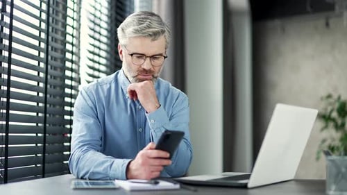 Thoughtful mature gray haired bearded businessman browsing smartphone sitting at workplace