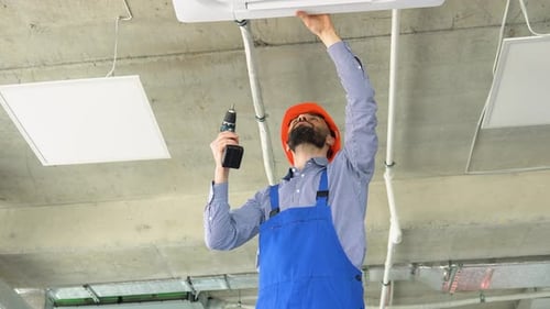 Young Worker in Uniform and Helmet Installs Air Conditioner in the Office