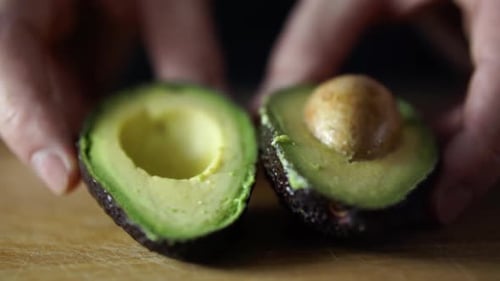 Mains d'un homme de race blanche poussant un avocat vert coupé en deux vers la caméra sur une planche à découper